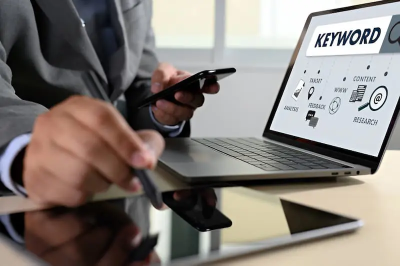 Person using a smartphone and stylus, with a laptop displaying a "Keyword" sign and icons.